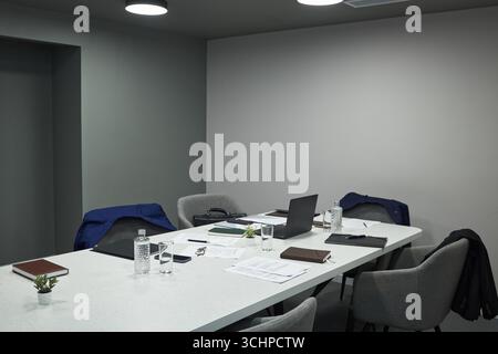 Moderner Büro-Besprechungsraum mit leeren Stühlen um den Konferenztisch, Anzugsjacken, offenem Laptop, Dokumenten, Notizbüchern, Wasserflaschen, Smartphone, kleine Pflanze, keine Leute anwesend Stockfoto
