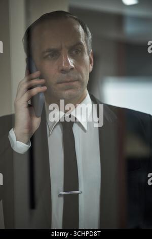 Kaukasier mittleren Alters, der drinnen steht und mit formellem Anzug und Krawatte auf dem Smartphone spricht, ernsthaft und konzentriert aussieht, Telefon am Ohr hält, kurzes Haar sichtbar, hinter einer Glaswand Stockfoto