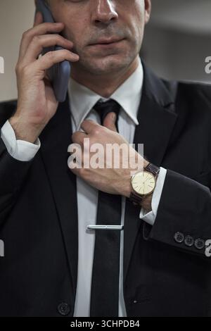 Anonymer Geschäftsmann passt die Krawatte an, während er mit dem Smartphone spricht, trägt formellen Anzug und Armbanduhr, partielles Gesicht sichtbar, Business Professional Setting impliziert Stockfoto