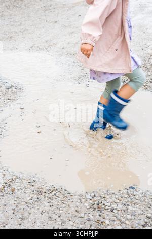 Kind mit Regenstiefeln, das in einer schlammigen Pfütze auf einem Kiesweg im Freien tritt Stockfoto
