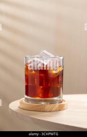Ein Glas eiskalten Americano-Kaffee mit Kristall-Eiswürfeln auf einem Holztisch, perfekt für Kaffeeliebhaber, die einen erfrischenden Moment kalter Brühe suchen. Stockfoto