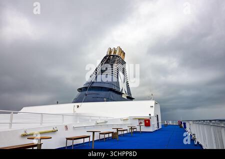 DFDS-Fährschornstein auf Øresund bei Rydebäck Schweden // RYDEBÄCK, Schweden — der Schornstein einer DFDS-Fähre ist vor einem bewölkten Himmel mit der Öresundstraße im Hintergrund sichtbar. Die Fähre trägt ein großes weißes maltesisches Kreuz-Emblem auf ihrem dunkelblauen Überbau. Das Deck der Fähre ist mit blauem Teppich, weißen Geländern und mehreren Holzbänken und Tischen ausgestattet. DFDS ist ein Dänisch-norwegisches Fährunternehmen, das Strecken durch Nordeuropa betreibt, einschließlich Verbindungen in der Ostsee und im Ärmelkanal. Die Routen des Unternehmens verbinden verschiedene Länder wie Dänemark, Norwegen und Swe Stockfoto