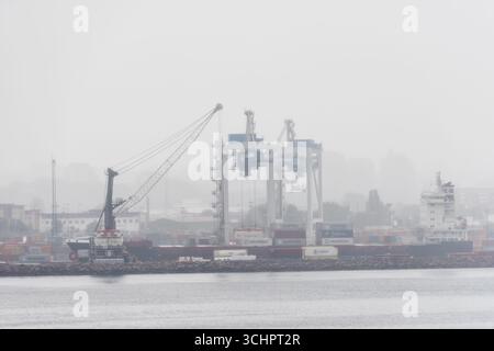 Containerschiffe und Krane im Hafen von Helsingborg Schweden in Nebel // HELSINGBORG, Schweden — Ein Containerschiff und Kräne sind im Hafen von Helsingborg, Schweden, bei Nebel und Nieselregen sichtbar. Der Hafen ist ein bedeutender logistischer Drehpunkt im Land, der den Güterverkehr über den Seeverkehr erleichtert. Die Szene erfasst die industrielle Aktivität des Hafens unter atmosphärischen Bedingungen, die die Sicht beeinträchtigen. Stockfoto