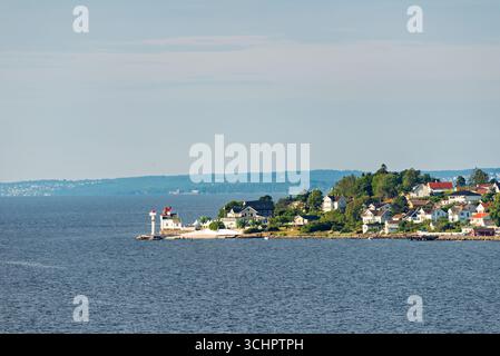 Leuchtturm in Ostensjovannet Vestby Norway // VESTBY, Norwegen — Ein Leuchtturm steht auf einer kleinen Halbinsel, die ins Wasser ragt, mit einer Ansammlung weißer Häuser und grüner Bäume, die das Ufer dahinter säumen. Der weiße Leuchtturm mit roten Akzenten ist ein markantes Merkmal der Küstenlandschaft. Die Szene fängt einen ruhigen Sommertag mit ruhigem Wasser und klarem Himmel ein und zeigt die malerische Landschaft von Vestby, einer Gemeinde in Akershus County, Norwegen. Die Gegend ist bekannt für ihre Schönheit an der Küste und ihre Wohngebiete, die sich mit der natürlichen Umgebung verbinden. Stockfoto