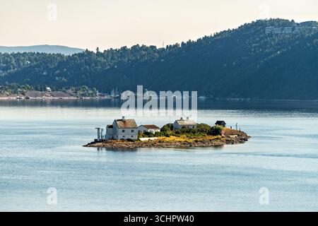 Steilene Fyr Leuchtturm Steilesand Norwegen / STEILESAND, Norwegen — der Leuchtturmkomplex Steilene Fyr befindet sich auf einer kleinen Insel vor der Küste in der Nähe von Steilesand in Akershus, Norwegen. Der Komplex umfasst ein primäres Leuchtturmgebäude und zugehörige Wohngebäude vor dem Hintergrund bewaldeter Hügel am Festlandufer, wobei sich das offene Wasser des Oslofjords zum Horizont hin erstreckt. Leuchttürme dieses Charakters sind charakteristisch für den inneren Fjord, wo die zahlreichen Inseln und die Schifffahrtsgefahren der am stärksten genutzten Küstenwasserstraße Norwegens ein ausgedehntes Netz von A erforderten Stockfoto