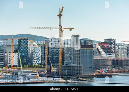 Baukrane über Ufergebäude Oslo Norwegen // OSLO, Norwegen — Baukrane über Ufergebäude in Oslo, Norwegen. Die Szene zeigt die anhaltende städtische Transformation des Hafengebietes der Stadt. Mehrere moderne Architekturstrukturen sind sichtbar, einige stehen kurz vor der Fertigstellung, andere befinden sich noch in frühen Bauphasen. Die Uferpromenade ist ein Zentrum der Aktivitäten, mit schweren Maschinen und Gerüsten, die auf bedeutende Entwicklungsprojekte hinweisen. Das Bild spiegelt das dynamische Wachstum und die Modernisierung des Stadtbildes Oslos entlang der Küste wider. Stockfoto