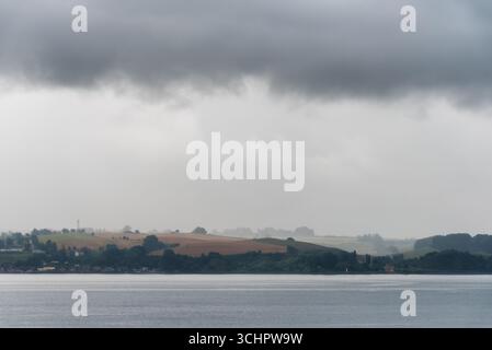 Küstenlandschaft mit Häusern und Hügeln Glumslöv Schweden / GLUMSLÖV, Schweden — Eine Küstenlandschaft mit sanften Hügeln und verstreuten Häusern entlang der Küste. Die Szene zeichnet sich durch einen Wasserkörper im Vordergrund und einen trüben, bewölkten Himmel aus. Glumslöv ist ein Dorf in der Gemeinde Landskrona in Skåne, Schweden, eine Region, die für ihre landwirtschaftlichen Flächen und die malerische Küstenlandschaft bekannt ist. Stockfoto