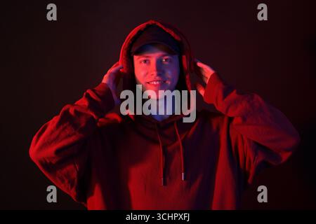 Porträt eines lächelnden Mannes mit Hoodie, Baseballkappe und Kopfhörern in Neonlichtern vor schwarzem Hintergrund Stockfoto