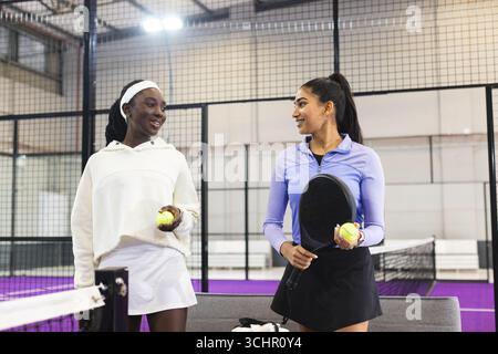 Verschiedene Freundinnen in Sportbekleidung stehen auf dem Padelplatz mit Schlägern und Tennisbälle am Netz Stockfoto