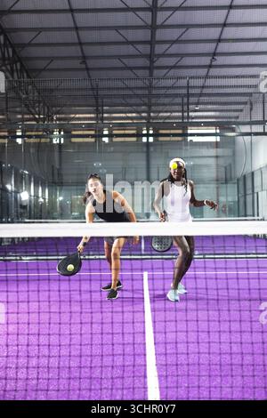Verschiedene weibliche Mannschaftskameraden schlagen auf dem Padel-Platz mit Schlägern einen gelb-grünen Ball über das Netz Stockfoto