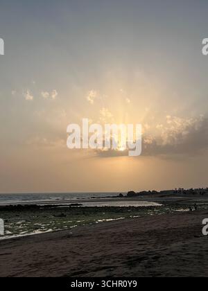 Sonnenuntergangslicht bricht durch die Wolken über dem Mokassar Beach mit felsiger Küste und Gezeitenreflexionen im südlichen Iran. Stockfoto