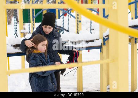 Zwei Kinder, ein Junge und ein Mädchen, sind auf einem schneebedeckten Spielplatz. Der Junge hilft seiner Schwester mit ihren Handschuhen. Sie werden im warmen Winter gebündelt Stockfoto