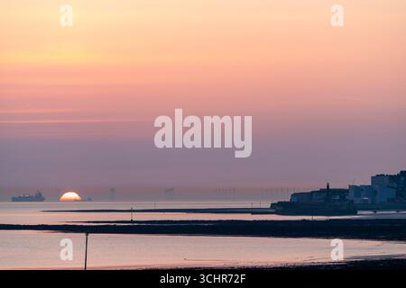 Sonnenaufgang über dem Meer und den Windturbinen des Thanet Offshore Windparks. Margate Harbour und die Wellenplattform bei Ebbe. Orangefarbener Himmel. Stockfoto