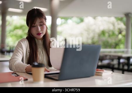 "Junge asiatische Frau konzentriert sich auf Laptops und Dokumente in einem produktiven freiberuflichen Arbeitsumfeld unter freiem Himmel" Stockfoto