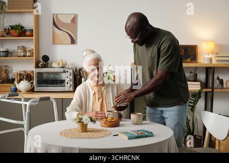 Ältere kaukasische Frau sitzt am Tisch mit einem Walker in der Nähe, Schwarzer Mann gießt Tee in eine Tasse, beide engagieren sich in freundlicher Interaktion in der häuslichen Küche, Buch und Snacks auf dem Tisch Stockfoto