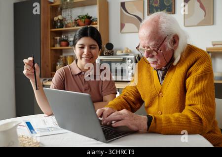 Ein älterer kaukasischer Mann benutzt Laptop, während eine junge Erwachsene hispanische Frau hilft und lächelt, beide sitzen mit Dokumenten am Tisch und arbeiten an digitalen Aufgaben im häuslichen Umfeld zusammen Stockfoto