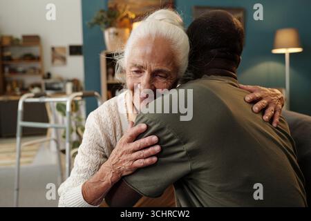 Ältere kaukasische Frau umarmt Schwarzen Mann im Wohnzimmer, beide zeigen Zuneigung und Wärme, ältere Frau lächelt mit geschlossenen Augen, Walker im Hintergrund sichtbar Stockfoto