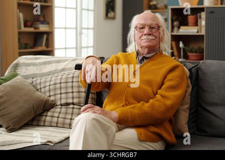 Porträt eines älteren kaukasischen Mannes, der auf dem Sofa sitzt und einen Gehstock mit der linken Hand hält, Brille trägt, mit ruhigem Ausdruck in die Kamera blickt, Haare und Schnurrbart sichtbar Stockfoto