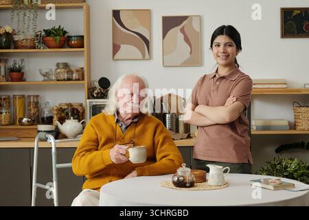Porträt eines älteren kaukasischen Mannes mit langen Haaren, der einen Becher hält, der am Tisch sitzt, mit einem Walker neben ihm, ein lächelndes Mädchen mit dunklen Haaren, das neben ihm steht und die Arme überkreuzt Stockfoto