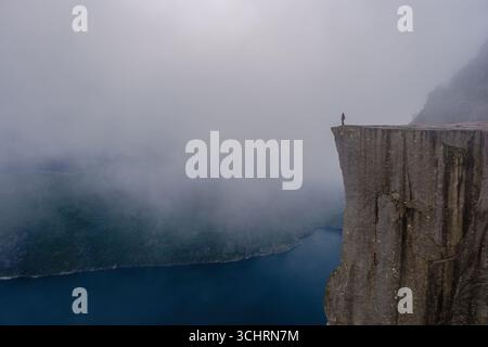 Majestätische Preikestolen Cliff steht hoch über dem Fjord, umgeben von Nebel. Eine einsame Figur genießt die spektakuläre Aussicht, die ein Gefühl von Abenteuer und Ruhe in der atemberaubenden Landschaft Norways weckt. Stockfoto