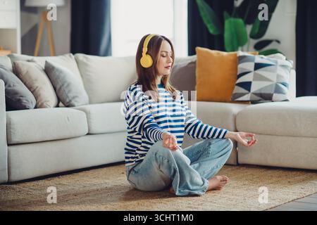 Junge Frau in gestreiftem Sweatshirt meditiert auf dem Wohnzimmerboden mit gelben Kopfhörern und genießt einen friedlichen Moment zu Hause Stockfoto