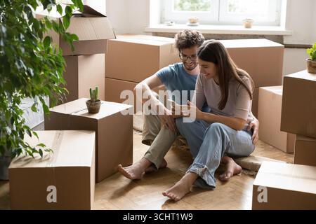 Glückliches Paar, das auf dem Boden zwischen Umzugskartons sitzt und das Telefon benutzt Stockfoto