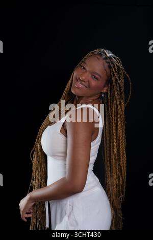 Schöne Frau mit geflochtenen Haaren, die lächelnd in die Kamera schaut, in einem weißen Outfit. Isoliert auf schwarzem Hintergrund. Fotoshooting. Stockfoto