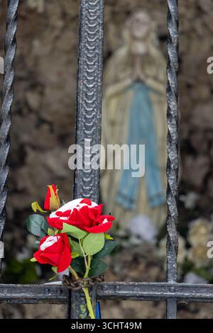 Künstliche rote Rosen, die mit Schnee bedeckt sind, sind an einem Eisenzaun befestigt, mit einer verschwommenen Marienstatue im Hintergrund. Stockfoto