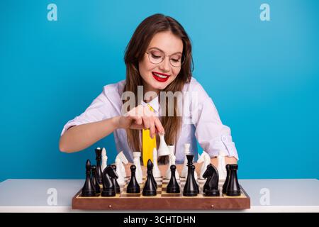 Kluge Frau, die Schach auf blauem Hintergrund spielt, strategisches Denken, akademische Ausrichtung und einen fröhlichen Ausdruck zeigt Stockfoto
