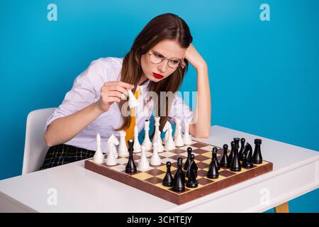 Junge Frau fokussiert, während sie Schach auf einem Tisch in einem hellblauen Raum spielt, um strategisches Denken und Konzentration zu erforschen Stockfoto