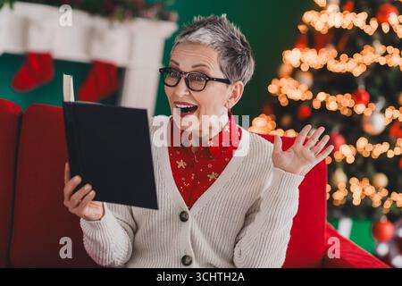 Ältere Frau genießt festliche Lektüre zu Hause am Weihnachtsbaum in gemütlicher, charmanter Urlaubsatmosphäre in Strickjacke Stockfoto