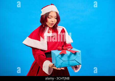 Junge Frau, die weihnachtsmann-Kostüm trägt und weihnachtsgeschenk hält, sieht überrascht auf blauem Hintergrund aus Stockfoto