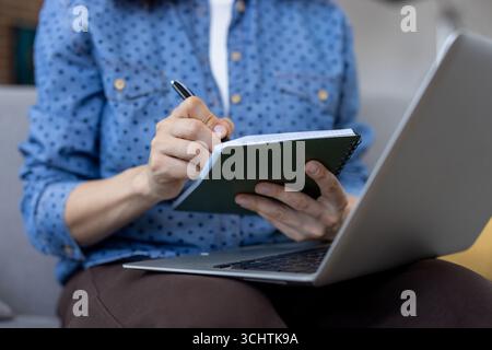 Eine Frau, die ein blaues Hemd trägt, macht Notizen in einem Notebook, während sie an einem Laptop arbeitet, auf einem Sofa im Heimbüro sitzt, und schlägt Fernarbeit oder Online-Studien vor Stockfoto