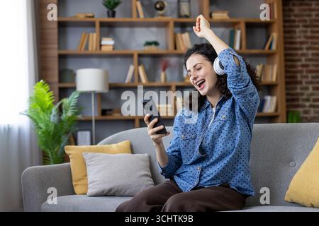 Junge Frau, die Kopfhörer trägt und Musik auf ihrem Smartphone hört, während sie auf einem bequemen Sofa in ihrem Wohnzimmer sitzt, ihre Lieblingslieder genießt und vor Freude tanzt Stockfoto