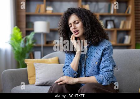 Junge Frau, die zu Hause auf dem Sofa sitzt, intensive Zahnschmerzen hat, ihre Hand gegen ihre Wange drückt und als Reaktion auf den überwältigenden Schmerz grimmig wird Stockfoto