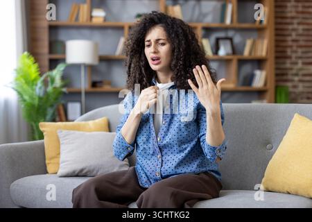 Junge Frau, die zu Hause auf dem Sofa sitzt und versucht, sich abzukühlen, leidet unter hohen Temperaturen und heißem Wetter, fühlt sich unwohl, fühlt sich unangenehm durch Hitzschlag im Sommer Stockfoto