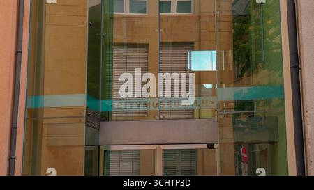LUXEMBURG, EUROPA - 1. SEPTEMBER 2025 - Glasfassade, die umliegende Gebäude mit venezianischen Jalousien und Bäumen reflektiert, Stadtmuseum Luxemburg, Luxemburg Stockfoto