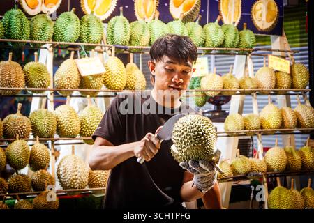 Ein junger asiatischer Mann schält geschickt einen Durianer mit einem Messer in einem lebhaften Obststand voller frischer Durianer jalan alor Food Street kuala lumpur Stockfoto
