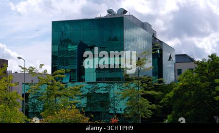 LUXEMBURG, EUROPA - 1. SEPTEMBER 2025 - grünes Glasgebäude mit bewölktem Himmel in Luxemburg-Stadt mit Bäumen und Straßenlaterne im Vordergrund Stockfoto