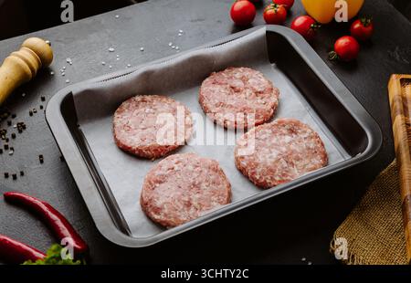 Frische, rohe Burgerkoteletts mit Gewürzen und Gemüse auf schwarzem Hintergrund, fertig zum Kochen Stockfoto