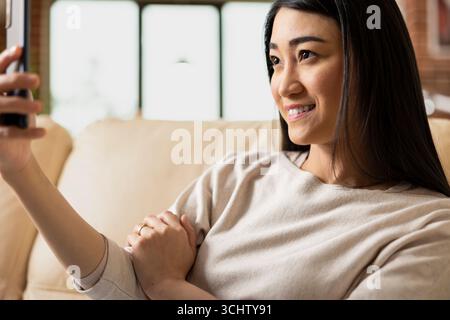 Lächelnde Unternehmerin sitzt bequem auf der Couch und macht Selfie mit dem Handy. Fröhlicher Remote-Mitarbeiter, der die persönliche Zeit genießt und den Tag für soziale Medien in einer gemauerten Wohnung dokumentiert. Stockfoto