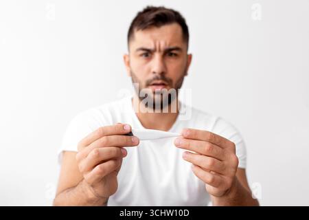 Trauriger junger Mann aus europa im weißen T-Shirt leidet unter gesundheitlichen Problemen, schaut auf Thermometer sitzt auf dem Bett Stockfoto