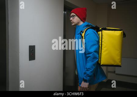 Der Liefermann mit blauer Jacke und rotem Hut wartet auf den Aufzug, während er einen gelben isolierten Thermorucksack für die Lieferung von Lebensmitteln trägt Stockfoto