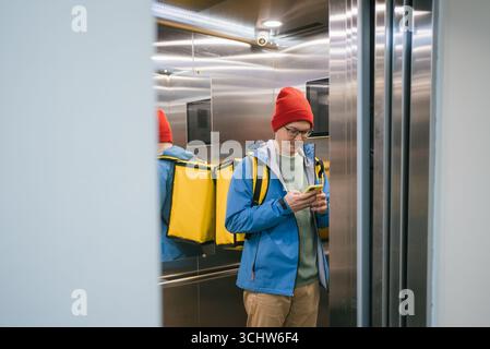 Der Liefermann mit rotem Wollhut und blauer Jacke überprüft sein Smartphone während der Fahrt in einem Aufzug und trägt einen gelben Thermorucksack für die Lieferung von Lebensmitteln Stockfoto