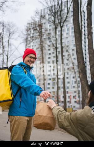 Mitarbeiter, der einen gelben Thermorucksack trägt, überreicht dem Empfänger eine Papiertüte mit Essen zum Mitnehmen in der Nähe von städtischen Grünflächen, was einen effizienten Service für die Lieferung von Lebensmitteln darstellt Stockfoto