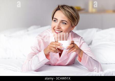 Glückliche Frau, die wegschaut und nachdenkt, Kaffee im Bett trinkt Stockfoto