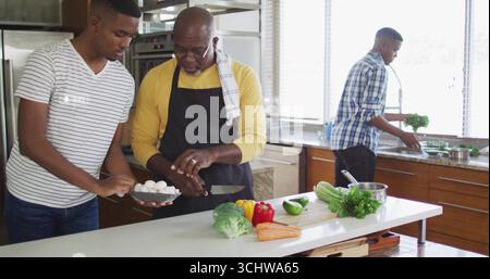 Er schneidet Vater in gelbes Hemd und Schürze, Sohn schneidet Pilze auf der Kücheninsel mit Gemüse Stockfoto