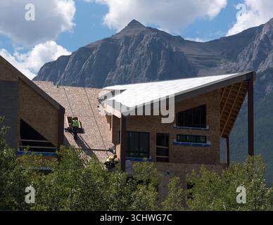 Zwei Bauarbeiter, die Kabelbäume verwenden, installieren Dachmaterialien in einem neuen Wohngebäude in Canmore, Alberta. Rundle Mountain bietet eine Kulisse. Stockfoto