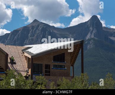 Zwei Bauarbeiter, die Kabelbäume verwenden, installieren Dachmaterialien in einem neuen Wohngebäude in Canmore, Alberta. Rundle Mountain bietet eine Kulisse. Stockfoto