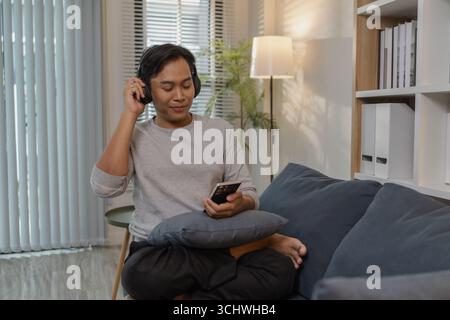 Ein junger Mann entspannt sich zu Hause und genießt Freizeit auf dem Sofa. Er hört Musik mit Kopfhörern, sieht Filme an und singt mit, expressi Stockfoto
